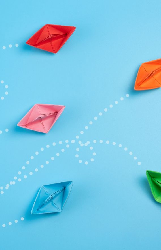 Bunte Papierboote sind auf einer blauen Oberfläche verstreut, wobei gepunktete weiße Linien skurrile, geschwungene Pfade bilden, die Bewegung oder Reisen suggerieren. Die Boote sind orange, rot, rosa, blau und grün.