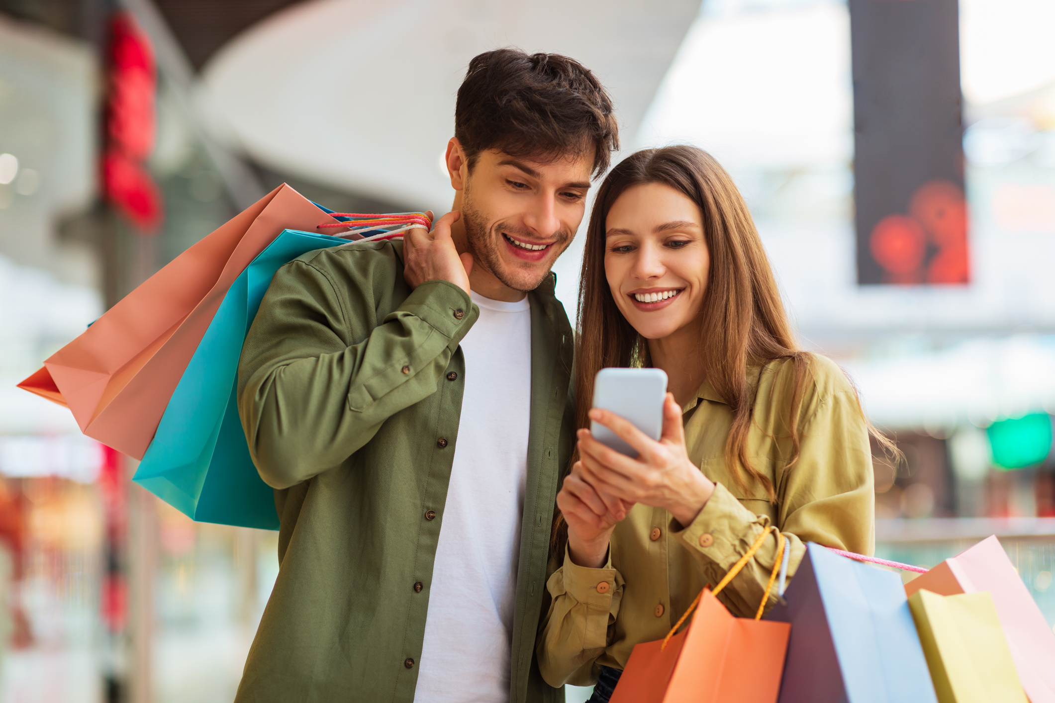 Lächelndes Pärchen beim Einkaufen, das bunte Taschen in der Hand hält und auf ein Smartphone in einem Einkaufszentrum schaut.