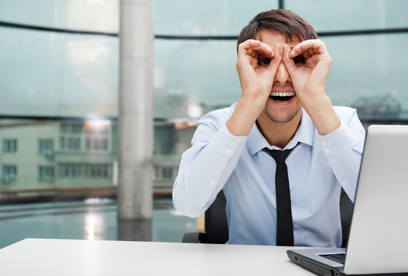 Lächelnder Mann in einem Büro, der mit seinen Händen ein Fernglas herstellt und an einem Schreibtisch mit einem Laptop sitzt.