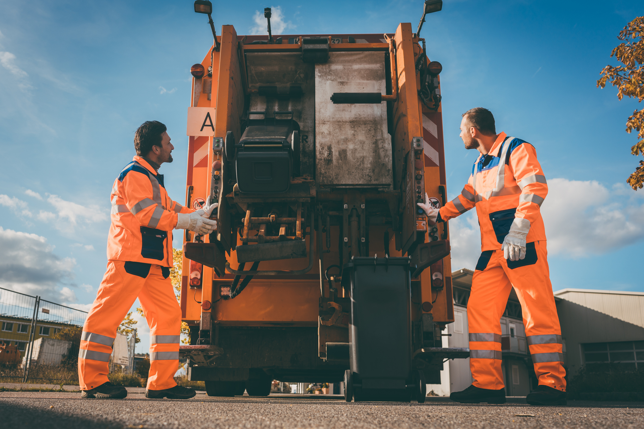 Zwei Arbeiter in orangefarbenen Uniformen laden an einem sonnigen Tag eine Mülltonne hinten in einen Müllwagen.