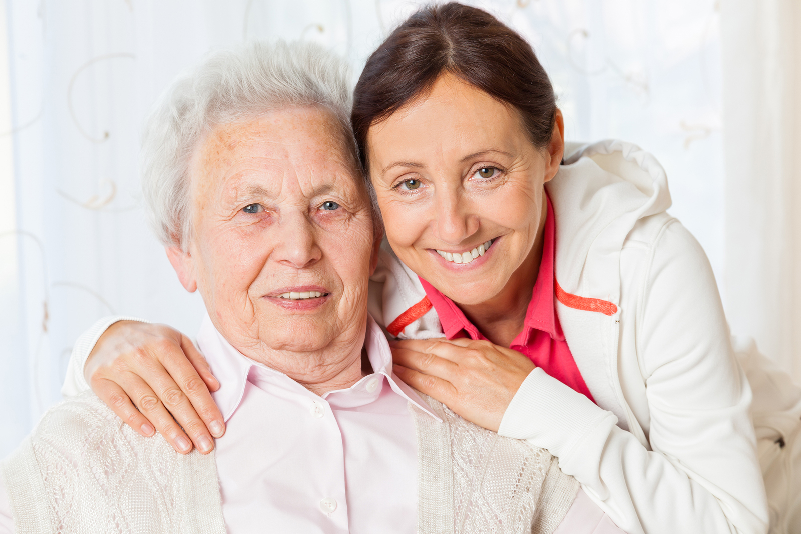 Zwei Frauen umarmen sich und lächeln in die Kamera. Die ältere Frau trägt Rosa, die jüngere eine weiße Jacke.