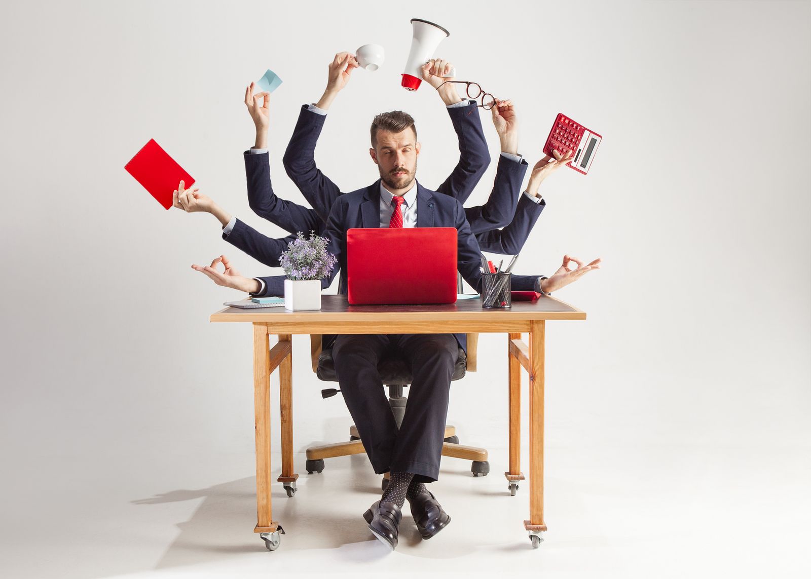 Ein Mann an einem Schreibtisch mit vielen Armen, der Multitasking betreibt, Büroartikel hält und an einem roten Laptop arbeitet.