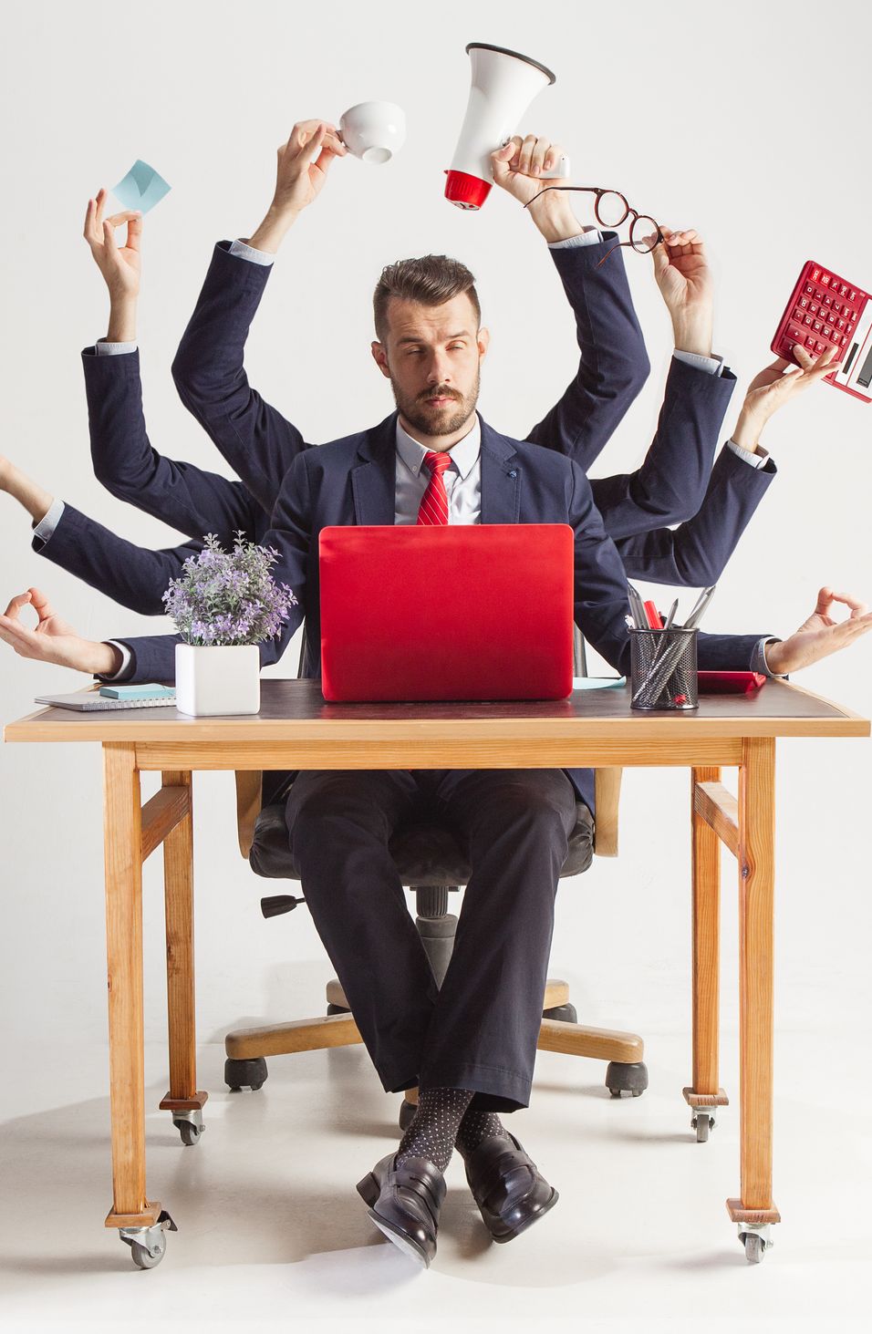 Ein Mann an einem Schreibtisch mit vielen Armen, der Multitasking betreibt, Büroartikel hält und an einem roten Laptop arbeitet.