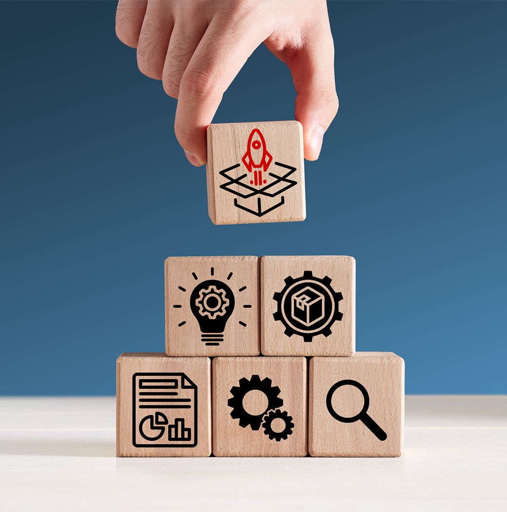 Eine Hand stapelt Holzblöcke mit den Symbolen einer Rakete, einer Glühbirne, eines Zahnrads, einer Tabelle, eines Würfels und einer Lupe.
