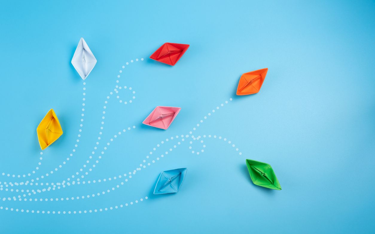 Bunte Papierboote sind auf einer blauen Oberfläche verstreut, wobei gepunktete weiße Linien skurrile, geschwungene Pfade bilden, die Bewegung oder Reisen suggerieren. Die Boote sind orange, rot, rosa, blau und grün.