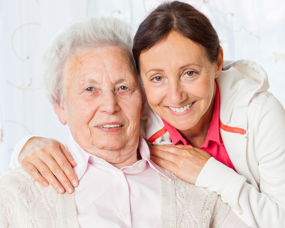 Zwei Frauen umarmen sich und lächeln in die Kamera. Die ältere Frau trägt Rosa, die jüngere eine weiße Jacke.