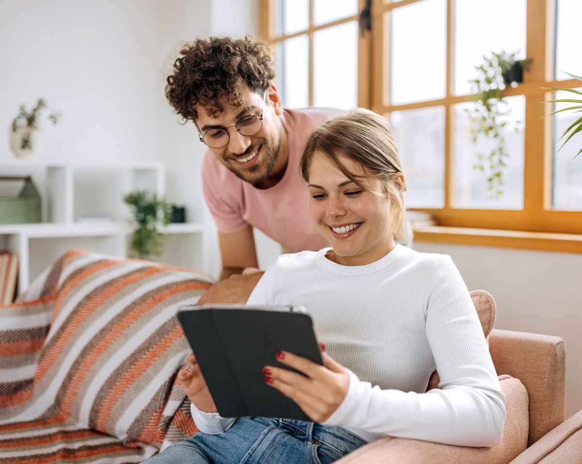 Eine lächelnde Frau benutzt ein Tablet, während ein Mann neben ihr steht. Beide wirken glücklich in einem gemütlichen Wohnzimmer.