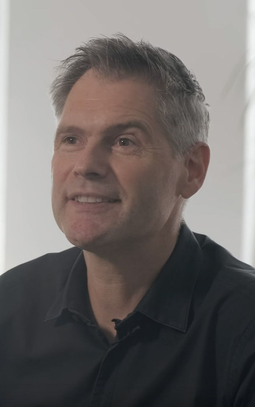 Ein Porträt von Michael Pietz welcher lächelt und leicht nach oben blickt. Er trägt ein dunkles Hemd und sitzt in einem hell erleuchteten Raum mit einem verschwommenen Hintergrund mit einem Fenster und hohen Pflanzen.