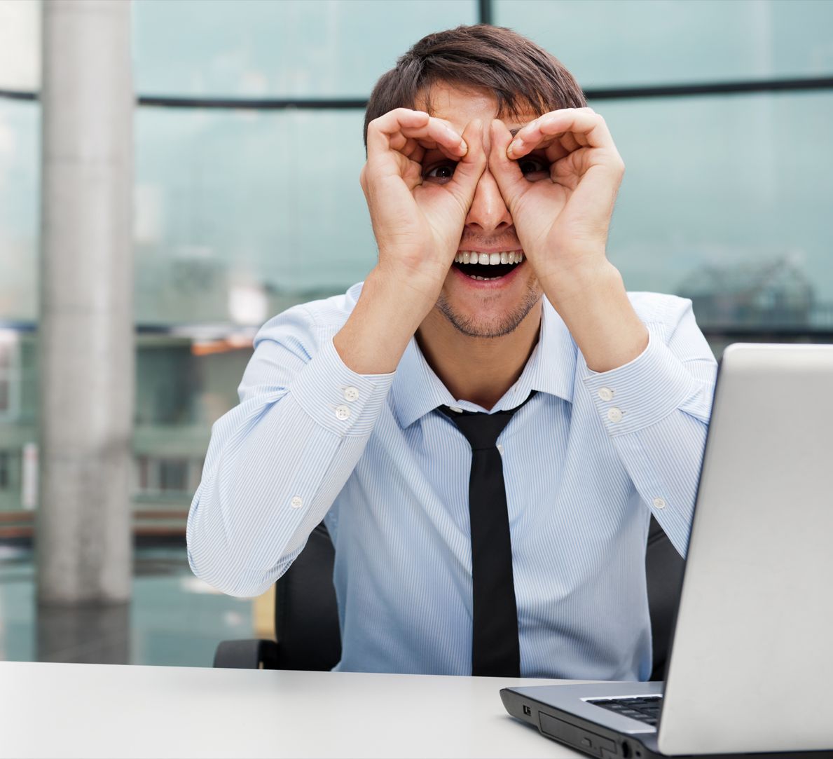 Lächelnder Mann in einem Büro, der mit seinen Händen ein Fernglas herstellt und an einem Schreibtisch mit einem Laptop sitzt.