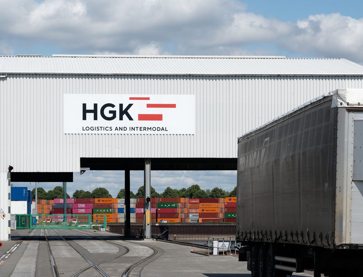 Ein Logistiklager mit HGK-Schild, einem Anhänger und Eisenbahnschienen, die zu bunten Frachtcontainern führen.